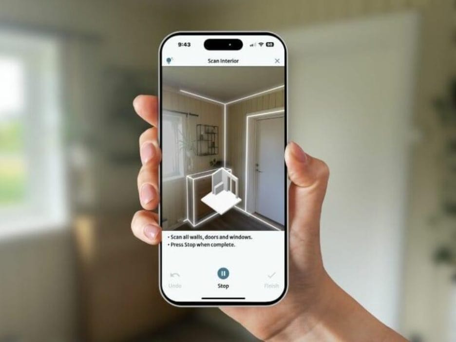 A hand is holding a smartphone running a room scanning application. The app interface shows an augmented reality overlay that outlines the edges of walls, doors, and windows in a room. On the screen, there’s a prompt instructing the user to “Scan all walls, doors and windows” and “Press Stop when complete.” The application appears to be capturing spatial data in real-time, using 3D visualization to generate an interior layout or floor plan. The interface also includes buttons for “Undo,” “Stop,” and “Finish,” indicating the scanning process is interactive and user-controlled.