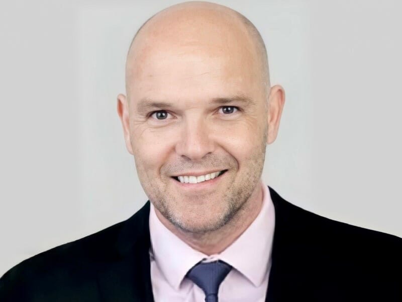 Real estate agent James Bellini in a suit and tie, wearing a light pink shirt and dark blazer, posing against a plain light background.