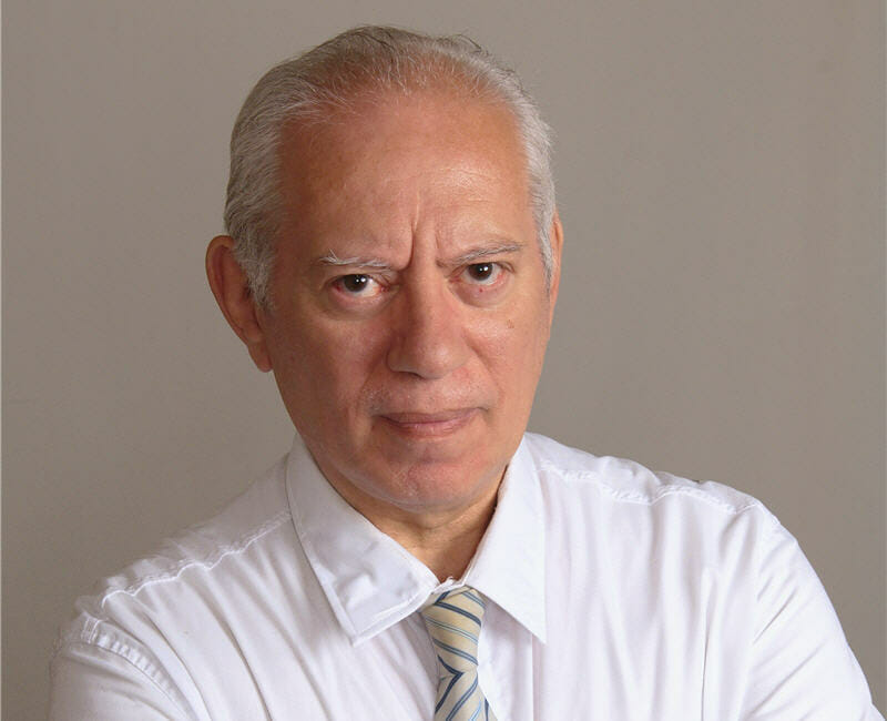 Architect Ruben Feldman wearing a white dress shirt and striped tie, looking at the camera with a serious expression against a plain background.